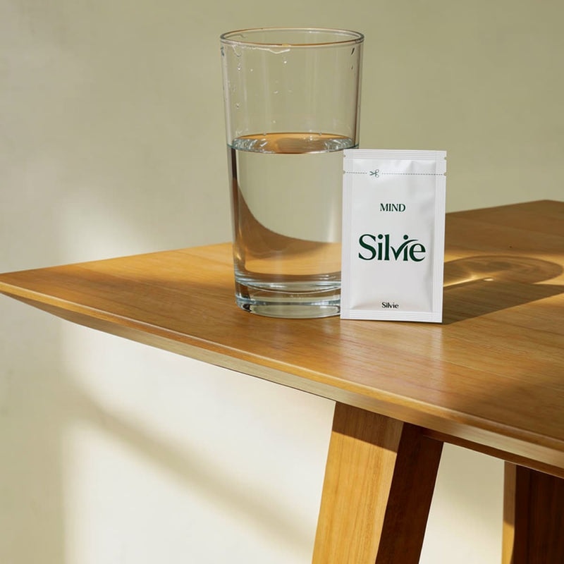 Clear glass of water and a white sachet of Silvie Mind on a wooden table.