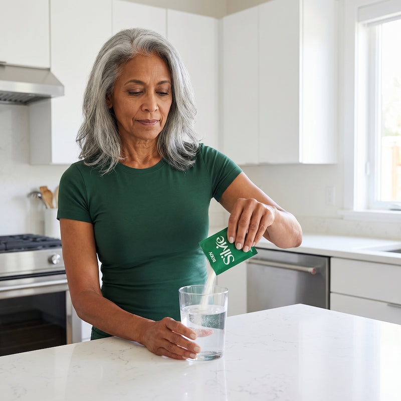 A middle-aged woman pouring supplement powder from Silvie supplement sachet into a glass of water