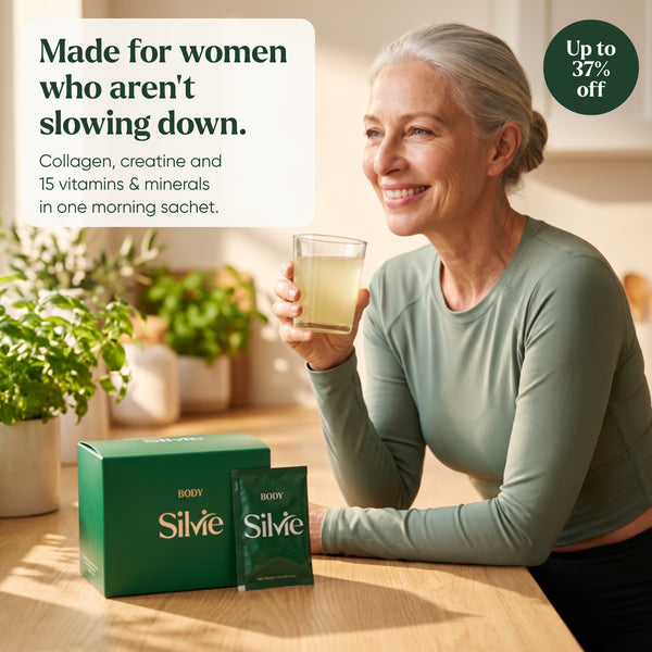 Woman in her 60s smiling with a glass of Silvie Body in a sunny kitchen, the box and a sachet on the table