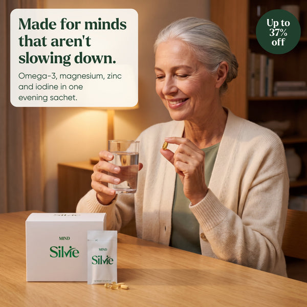 Woman in her 60s taking a Silvie Mind softgel with a glass of water in a warm living room, box and sachet on the table