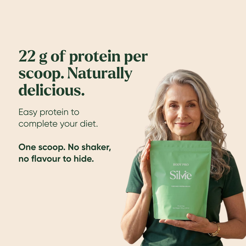 Woman in her 60s adding a scoop of Silvie Body Pro to a yoghurt bowl in a sunny kitchen, the pouch beside her